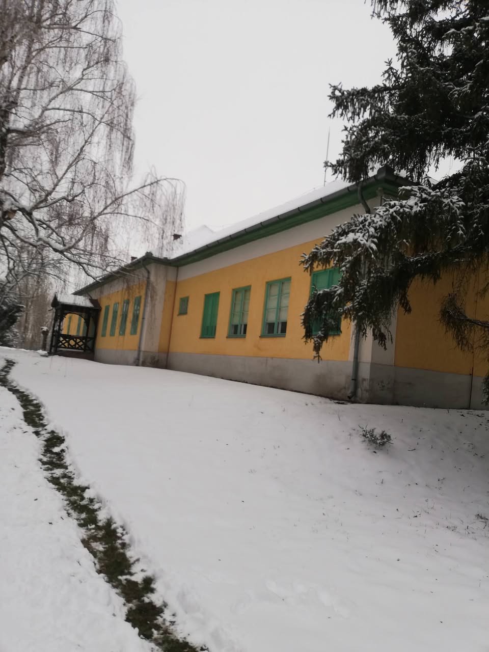 Ugyanaz a hosszú, sárga épület télen, hófödte udvarral és havas fenyőágakkal.