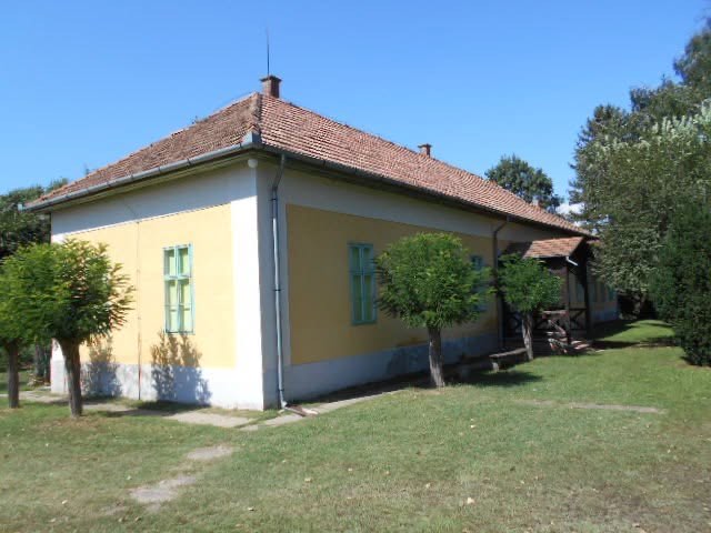 A vendégház sárga homlokzata zöld zsalugáterekkel, előtte nyírott gyep és fiatal fák.